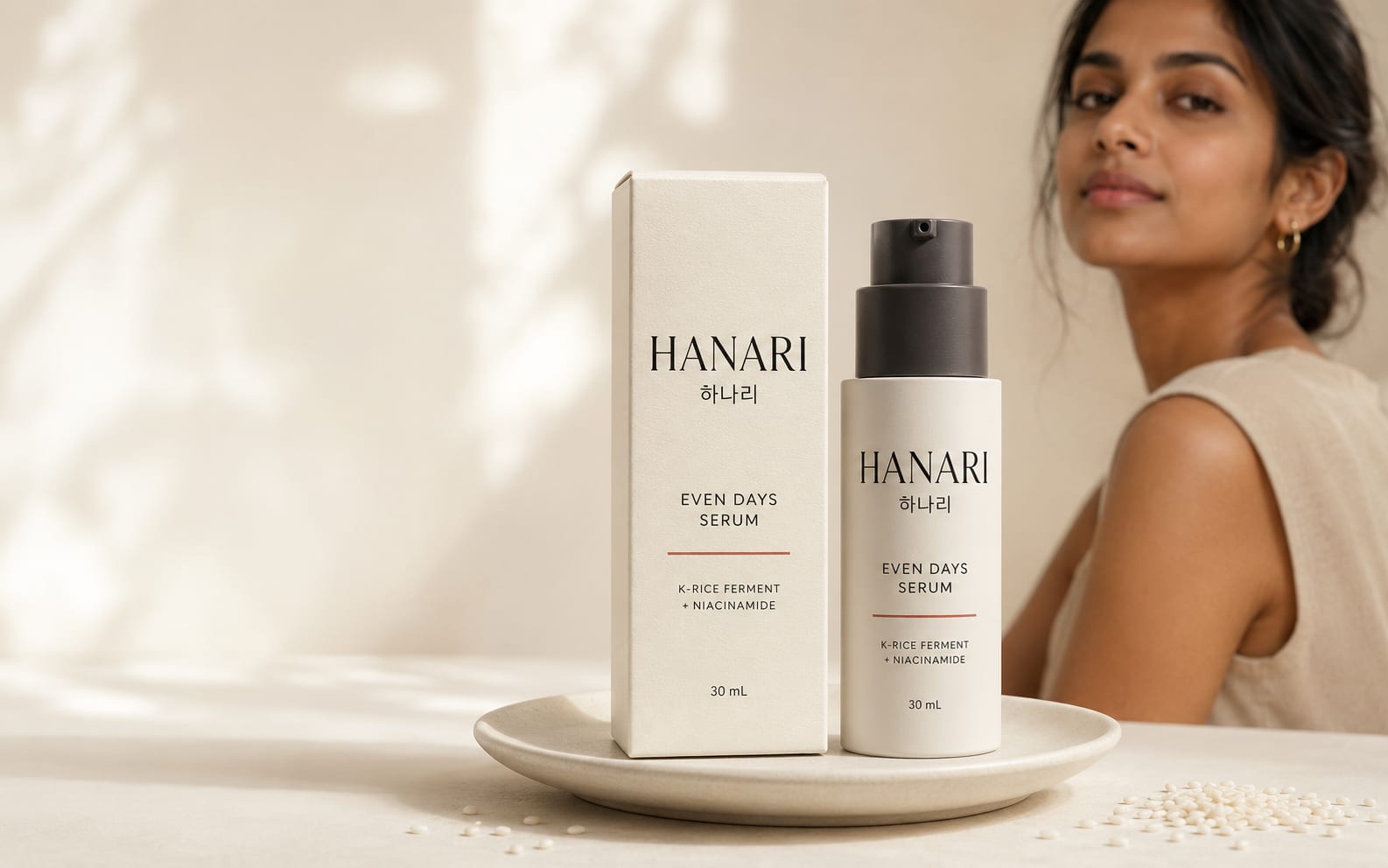 HANARI Even Days Serum and its carton on a ceramic plate, with rice grains scattered beside it.