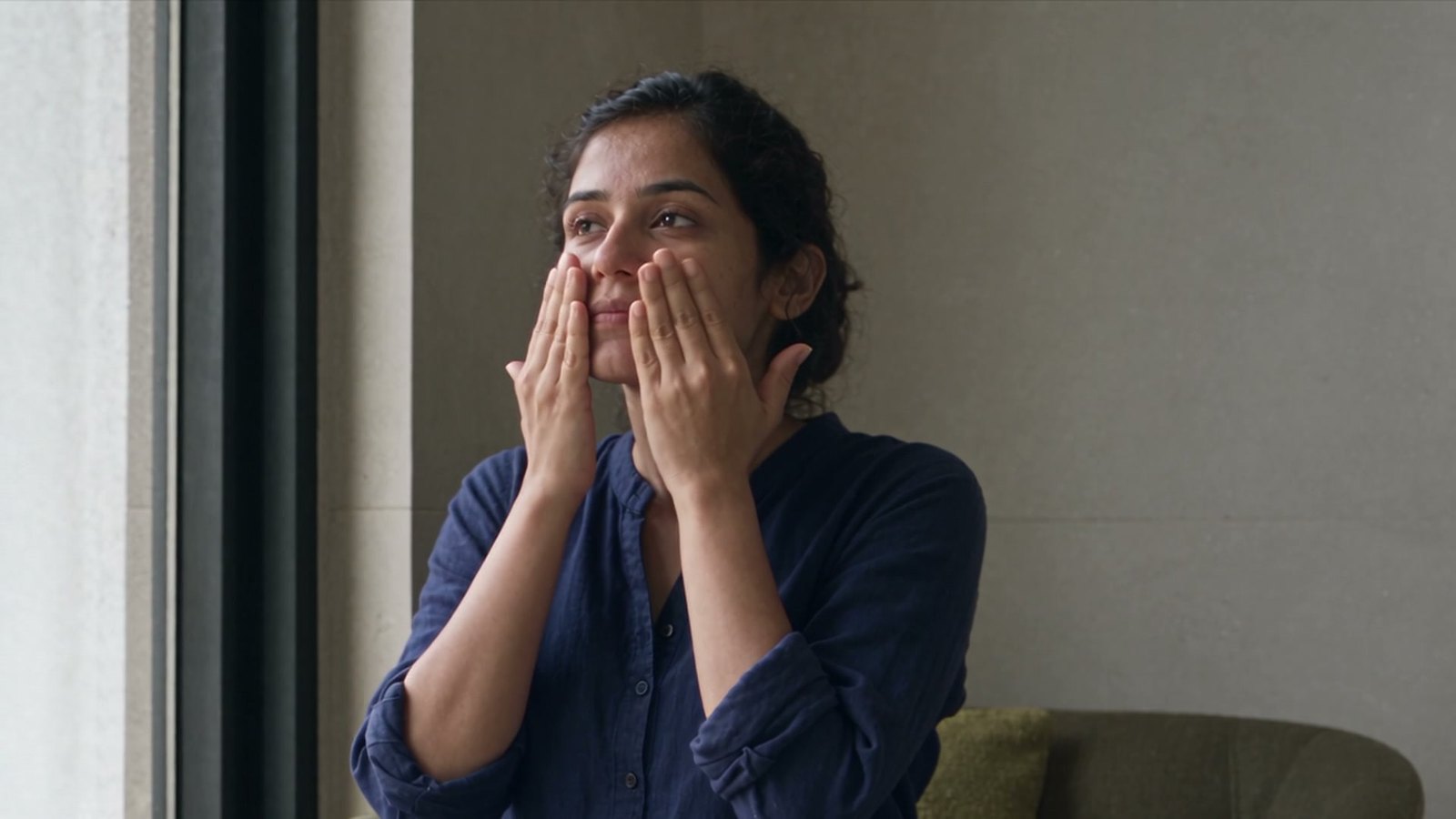 A woman by a window pressing both palms gently to her face